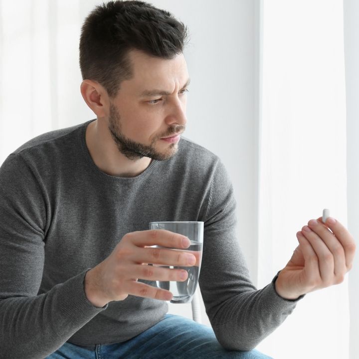 Man holding glass of water and examining medication pill.
